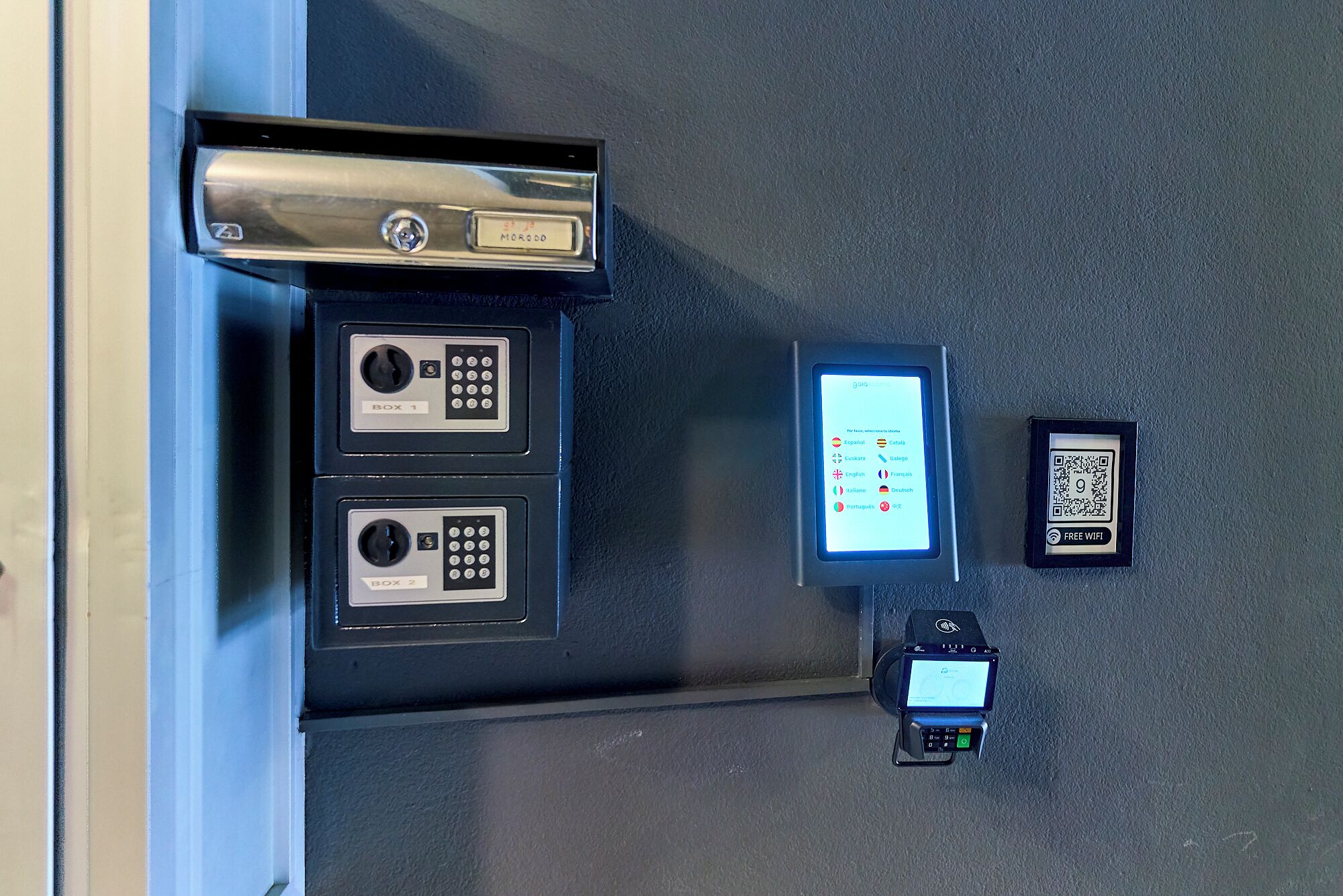Een zelfbedieningskiosk met een QR-code voor WiFi, een betaalterminal en twee genummerde vakken, waarschijnlijk voor toegangskaarten of sleutelkaarten.