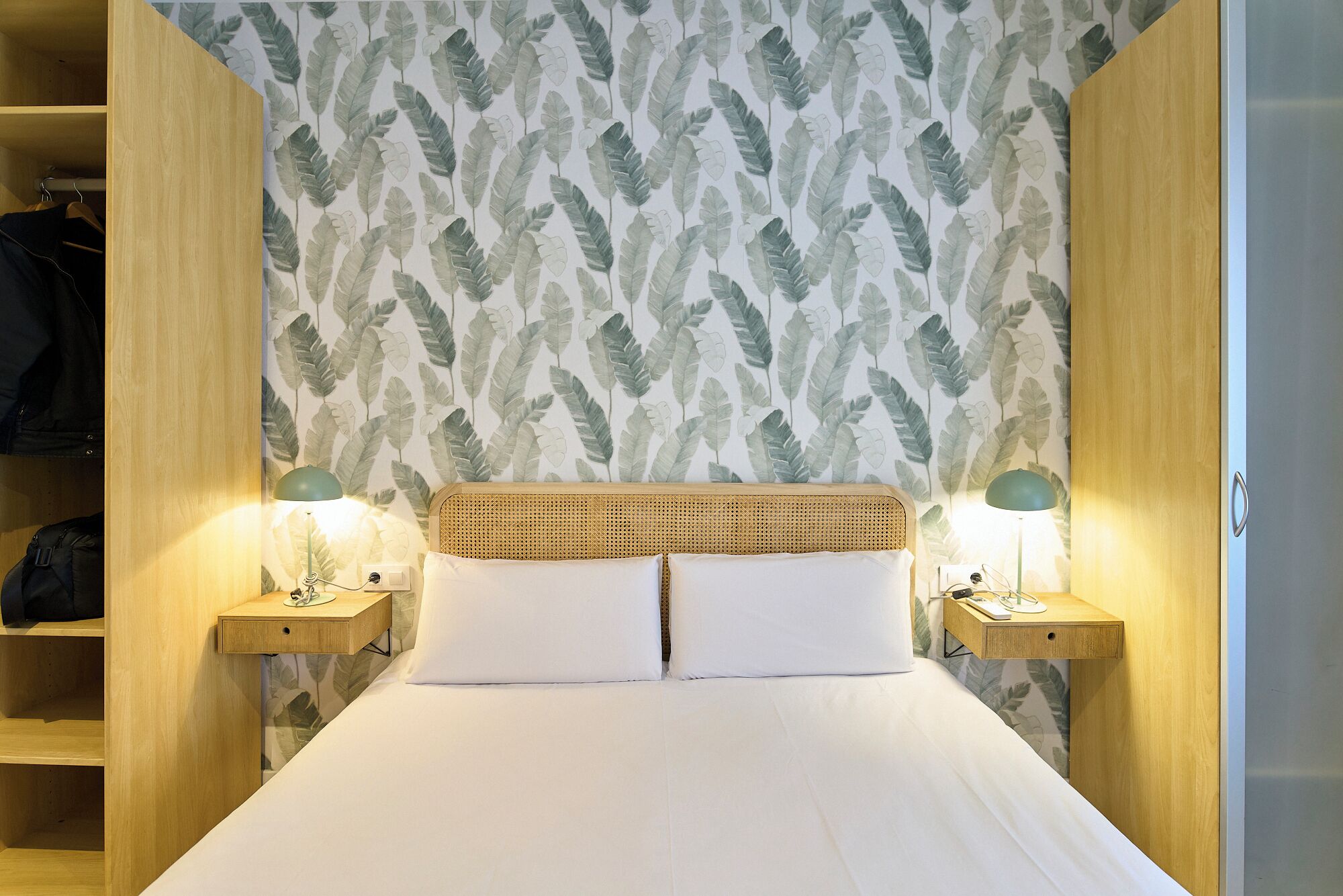 Een slaapkamer met een bed, twee nachtkastjes met lampen, een garderobe en een muur met een bladmotief.