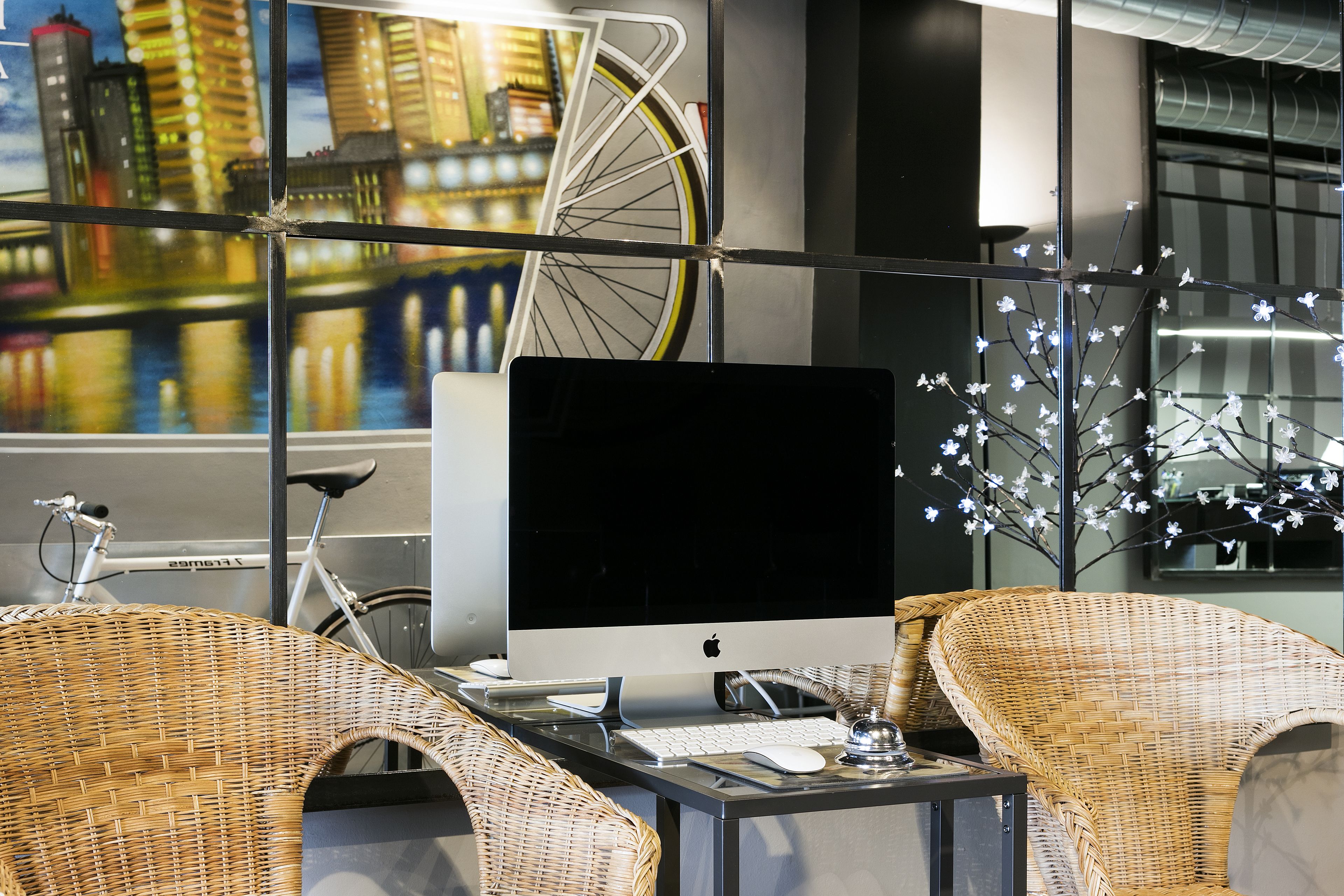 Un'area di lavoro moderna con un monitor, tastiera e mouse su un tavolo, circondata da poltrone in vimini. Una bicicletta è appesa al muro, e c'è un albero decorativo illuminato.