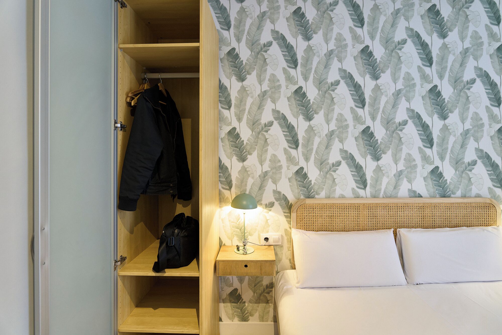 A bedroom with a bed, a nightstand with a lamp, a wardrobe with clothes and a bag, and a wall with a leaf pattern.
