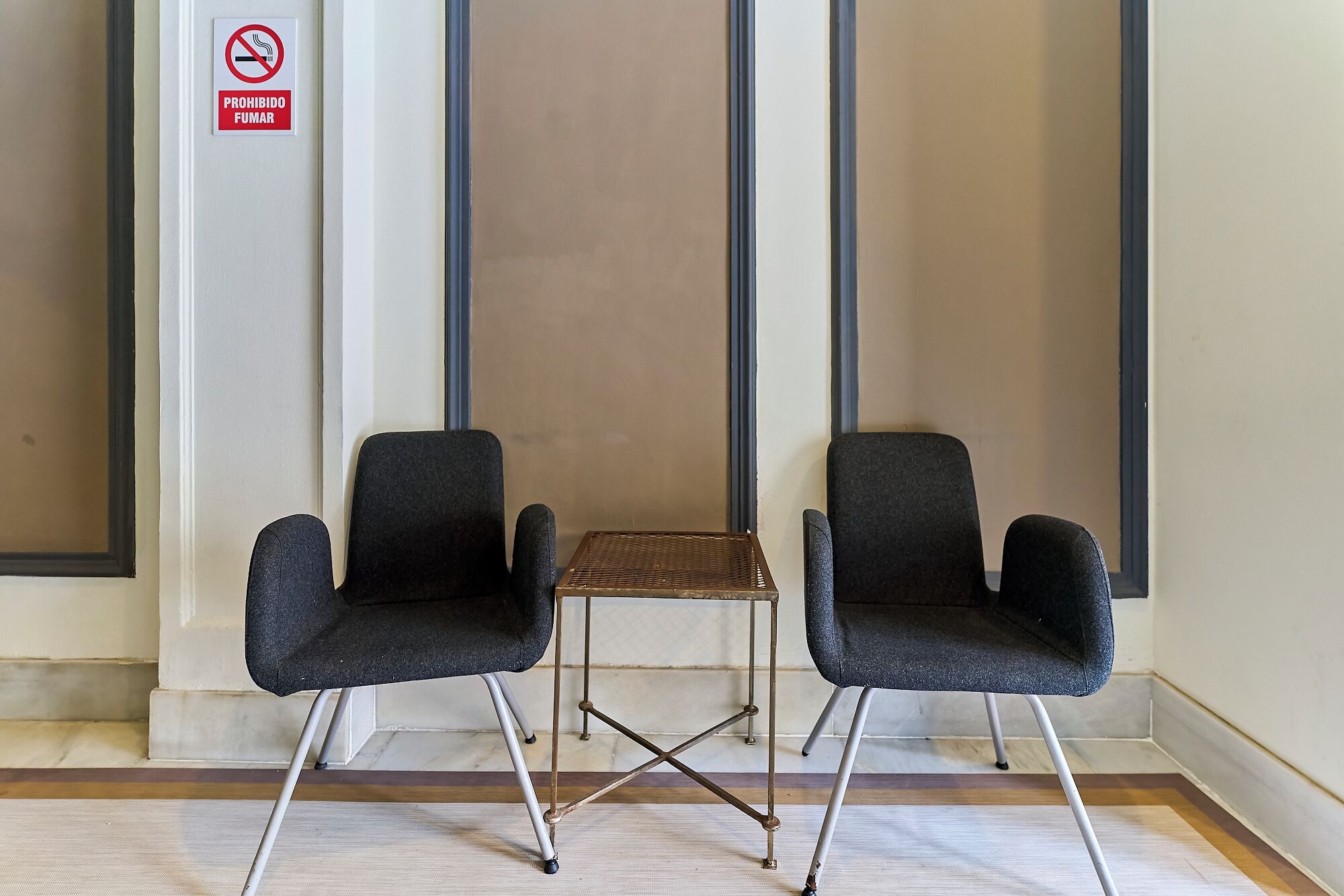 Two black chairs with metal legs and a small table in front of a wall with a 'No Smoking' sign.