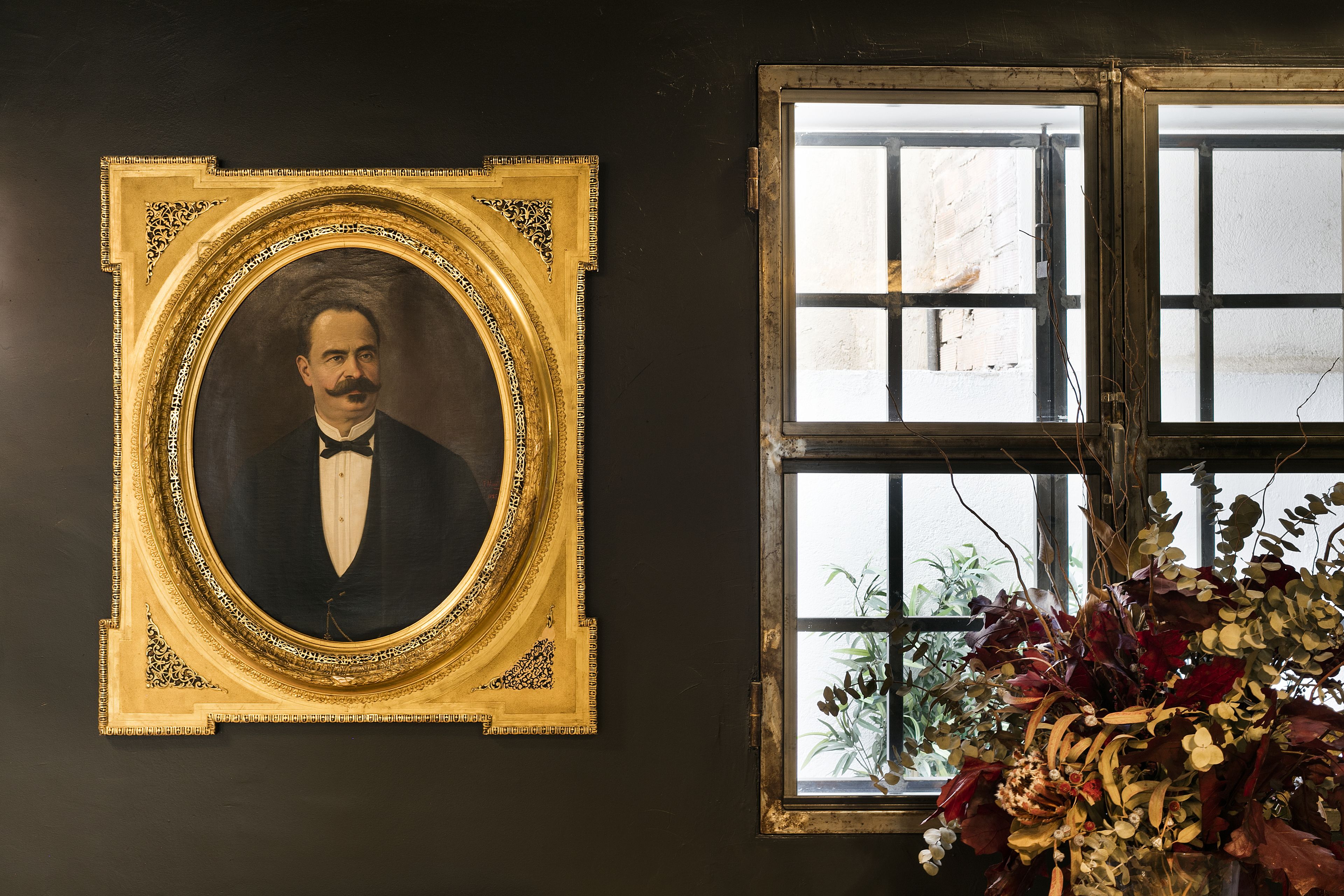Un retrato enmarcado en oro, una ventana con vistas al exterior y un jarrón con plantas secas.
