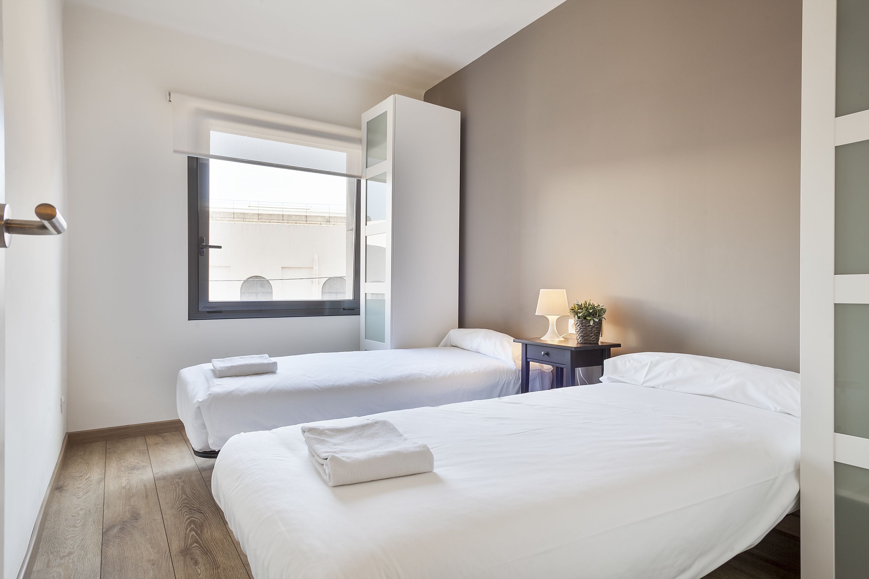 Habitación de hotel con dos camas, una ventana, una mesita de noche con lámpara y una pequeña planta.