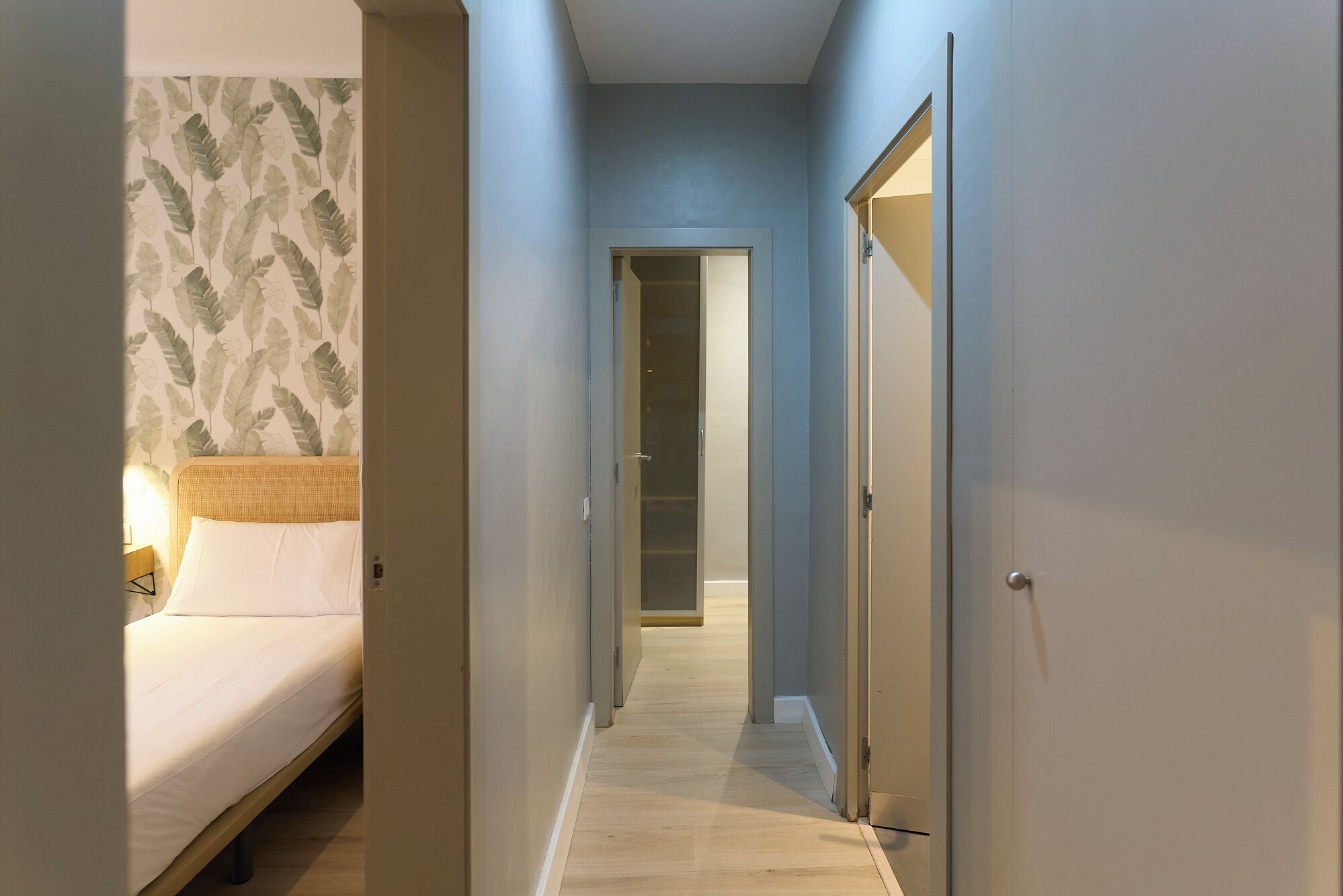 Un pasillo estrecho con una cama a la izquierda, una puerta a la derecha y otra al final.