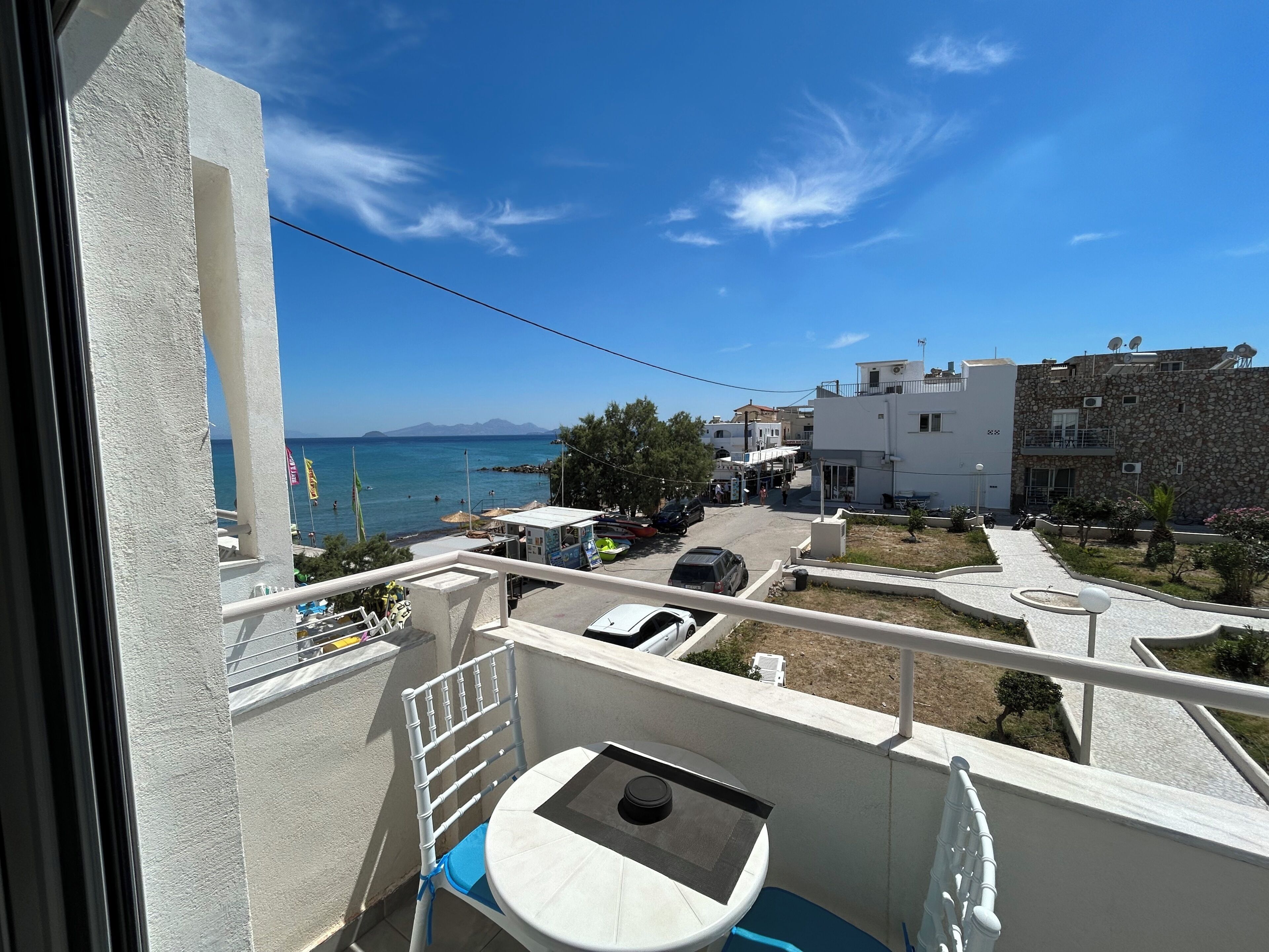 Ein Balkon mit Tisch und Stuhl, mit Blick auf eine Küstenlandschaft mit Gebäuden und geparkten Autos.