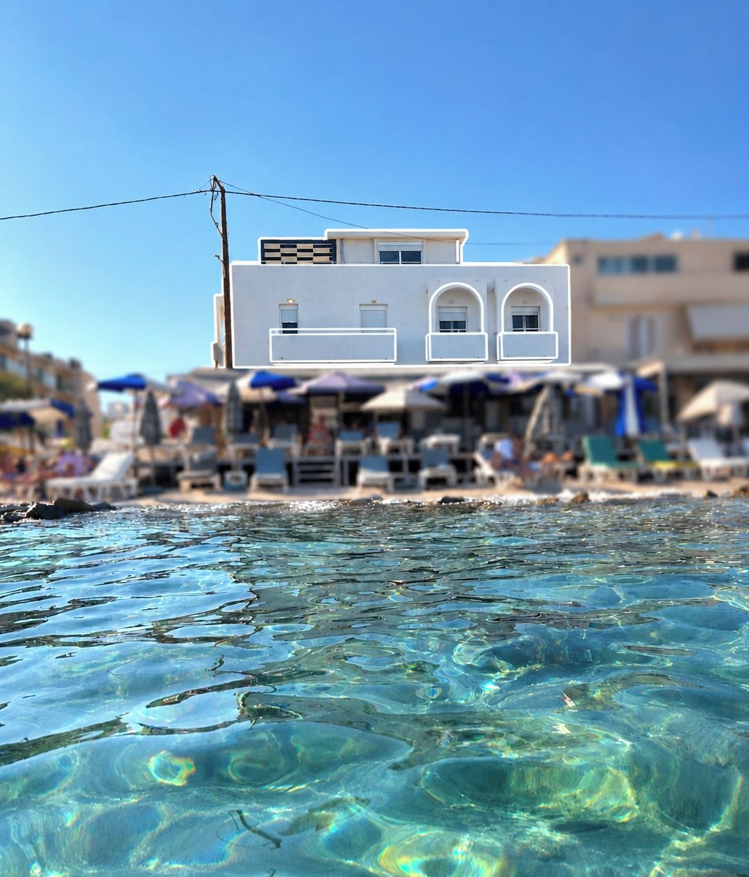 A beachfront hotel with clear blue water in the foreground, a white building with arched windows, and a beach area with umbrellas and lounge chairs.