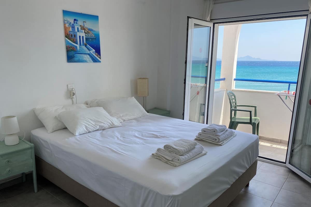Habitación de hotel con cama, un cuadro, una mesita de noche, una lámpara y vista al mar a través de una puerta de cristal.