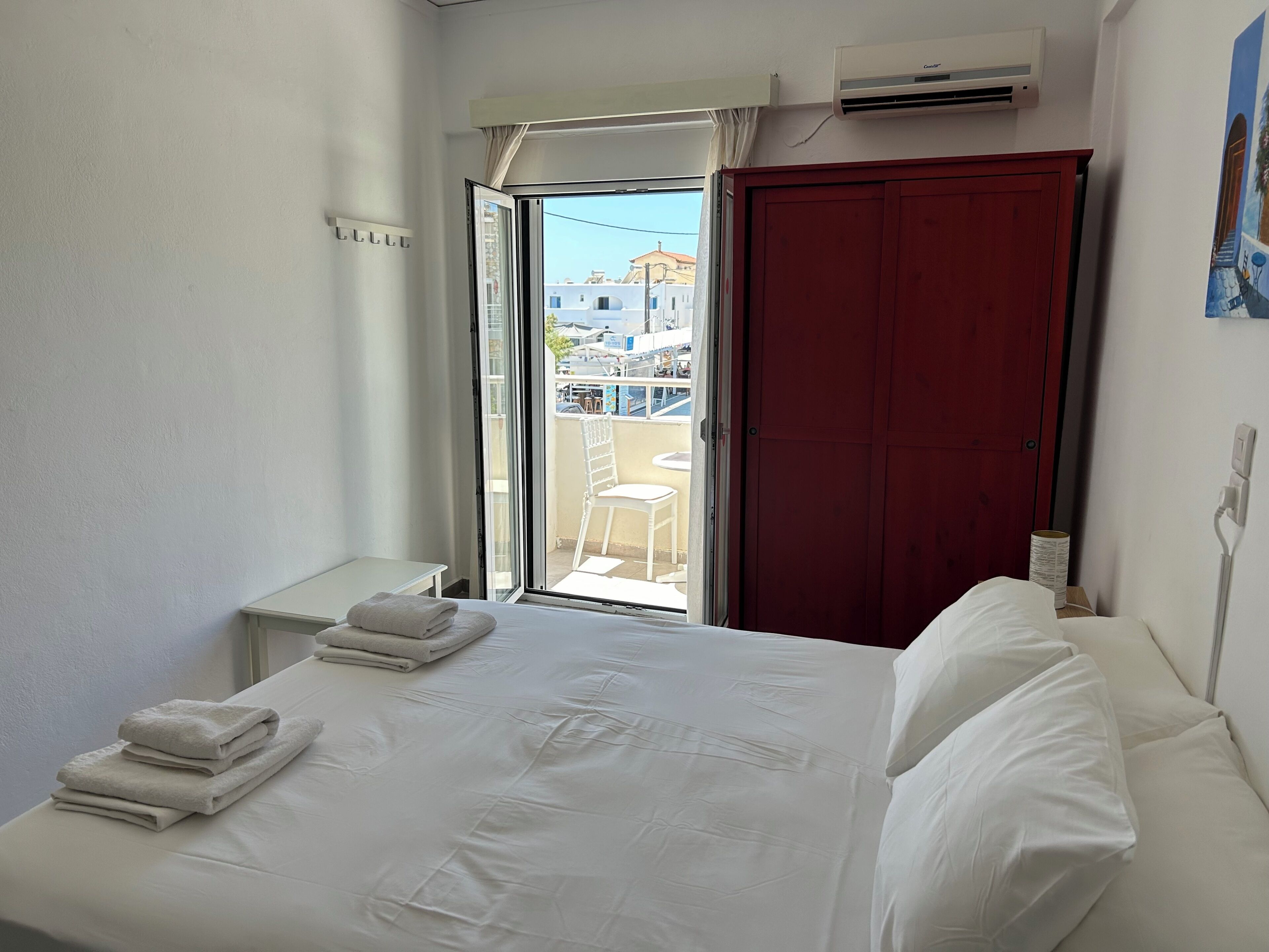A hotel room with a bed, a window with a view of buildings, a red door, and an air conditioning unit.