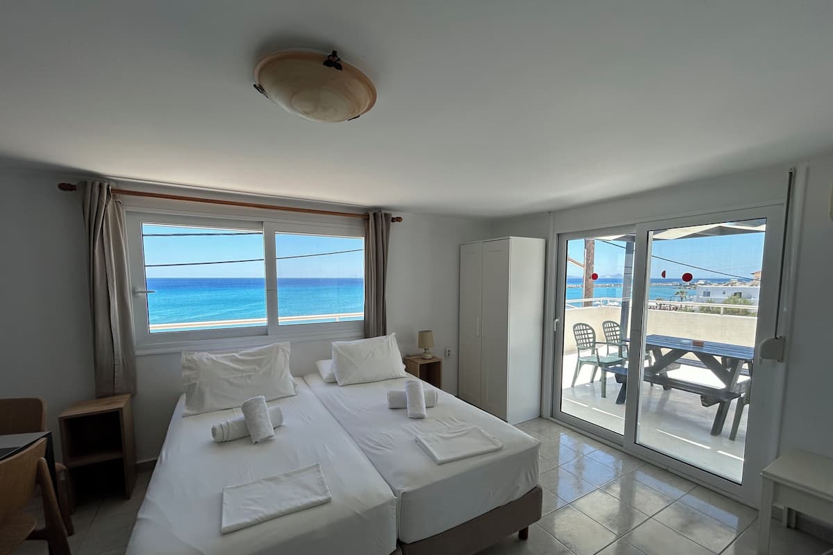 Habitación de hotel con dos camas, vista al mar, balcón con mesa y sillas, y puerta corrediza de vidrio.
