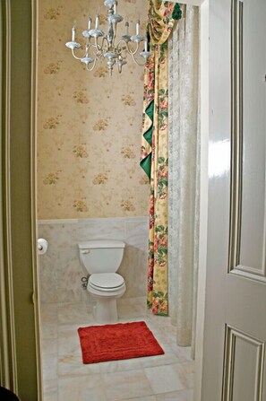 A bathroom with a toilet, a red rug, floral wallpaper, and a chandelier. - The Dansereau House (Thibodaux)