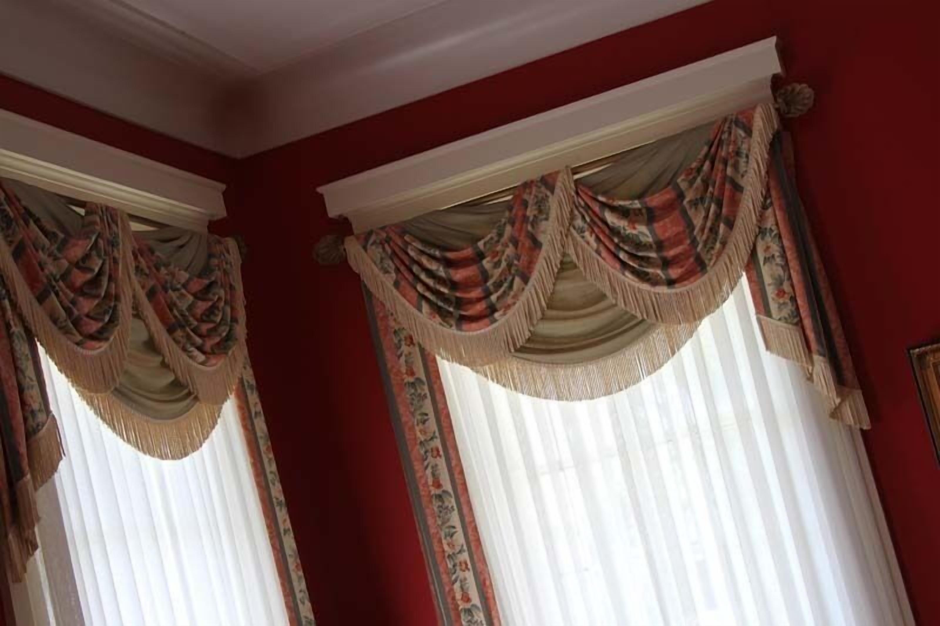 A room with red walls, two windows with white curtains, and decorative drapes with tassels.