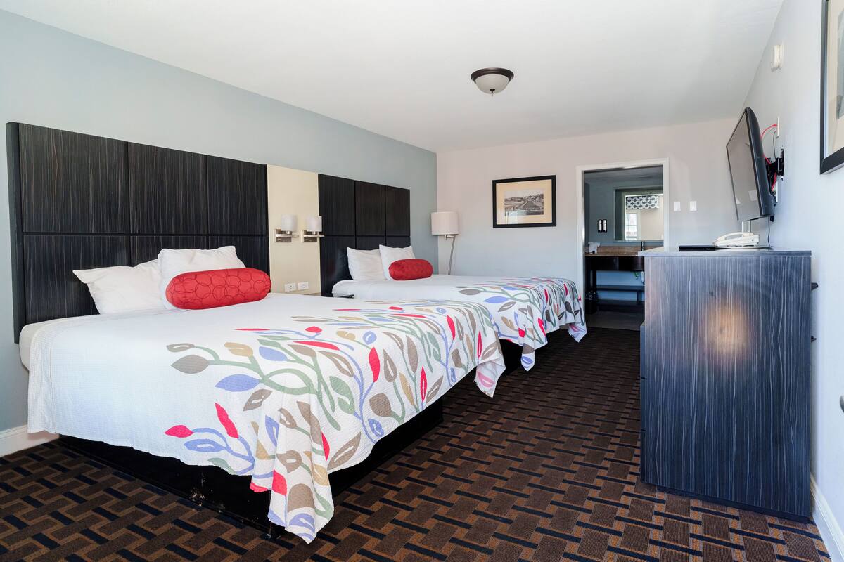 A hotel room with two beds, a dark headboard, a patterned bedspread, a wooden floor, a desk with a computer, and a framed picture on the wall.