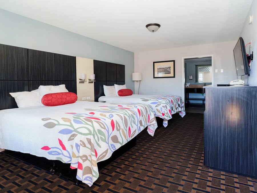 Habitación de hotel con dos camas, cabecera oscura, colcha estampada, suelo de madera, escritorio con computadora y un cuadro en la pared.
