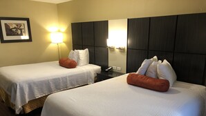 Deux lits simples avec du linge de lit blanc et des coussins décoratifs orange, une table de chevet avec un téléphone et un luminaire fixé au mur.