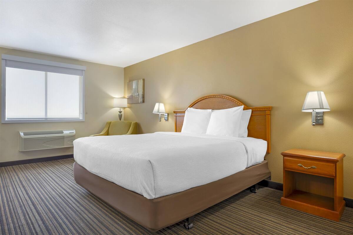 A hotel room with a large bed, two bedside lamps, a wooden nightstand, a window with blinds, and a wall-mounted air conditioner.