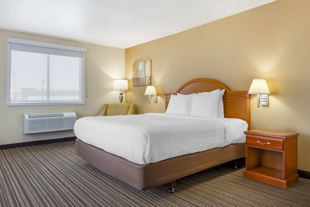 A hotel room with a large bed, two bedside lamps, a nightstand, a window with blinds, and a wall-mounted air conditioner.
