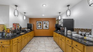 A modern kitchen with wooden cabinets, black countertops, and two pendant lights. There are framed pictures on the wall and a coffee machine on the counter. - Best Western East El Paso Inn (Clint)