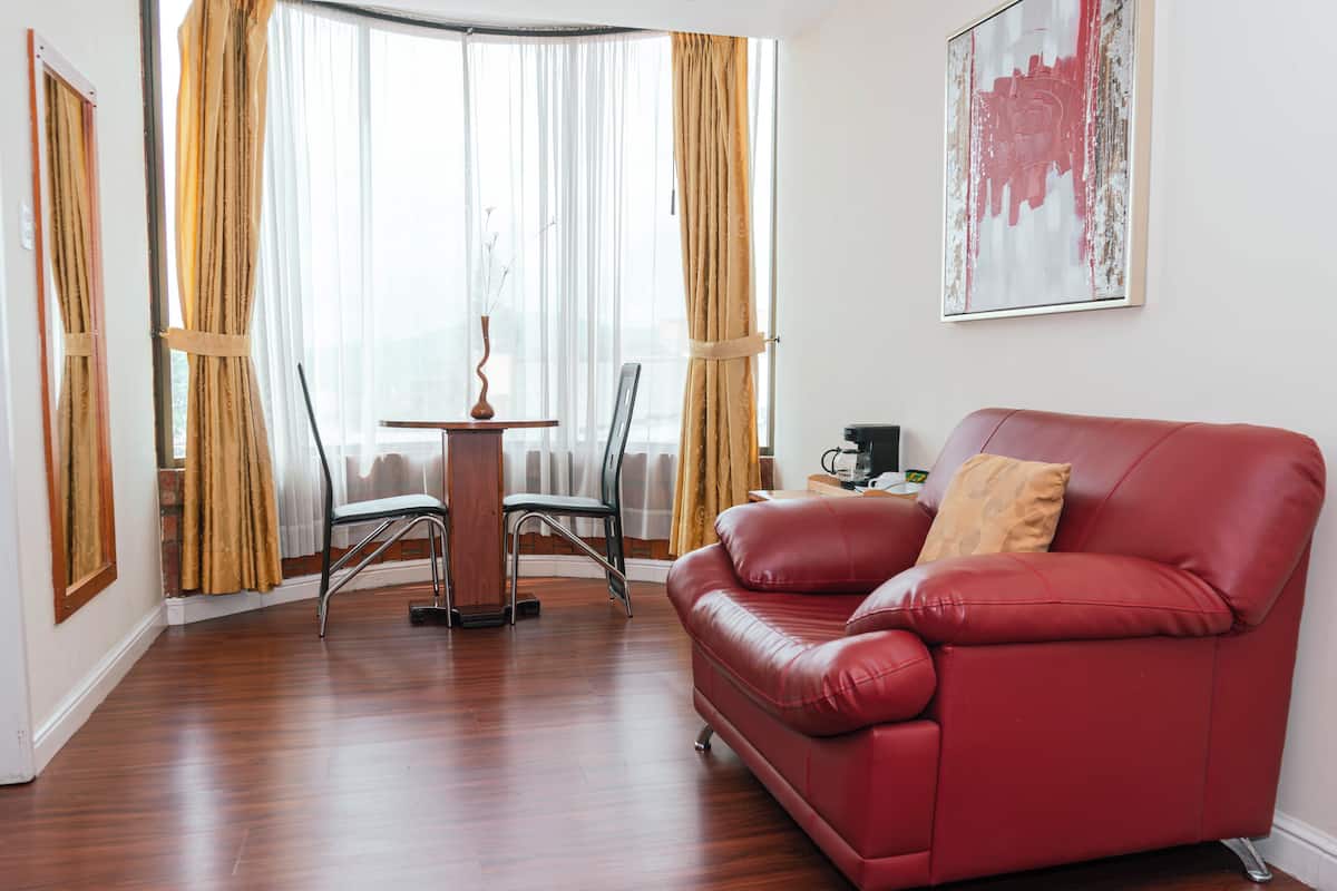 Un sofá de cuero rojo, una mesa de comedor con sillas y una cafetera en una habitación con suelo de madera y ventanas grandes con cortinas.