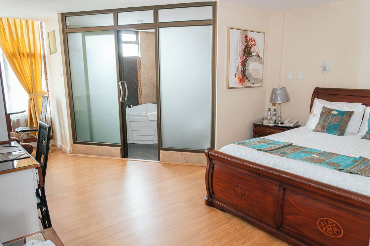 Habitación de hotel con cama de madera, un escritorio con silla, baño con bañera y puerta de vidrio corredera.