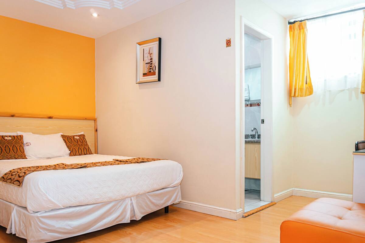 Una habitación de hotel con una cama, una pequeña silla naranja, una puerta que da a un baño y un cuadro en la pared.