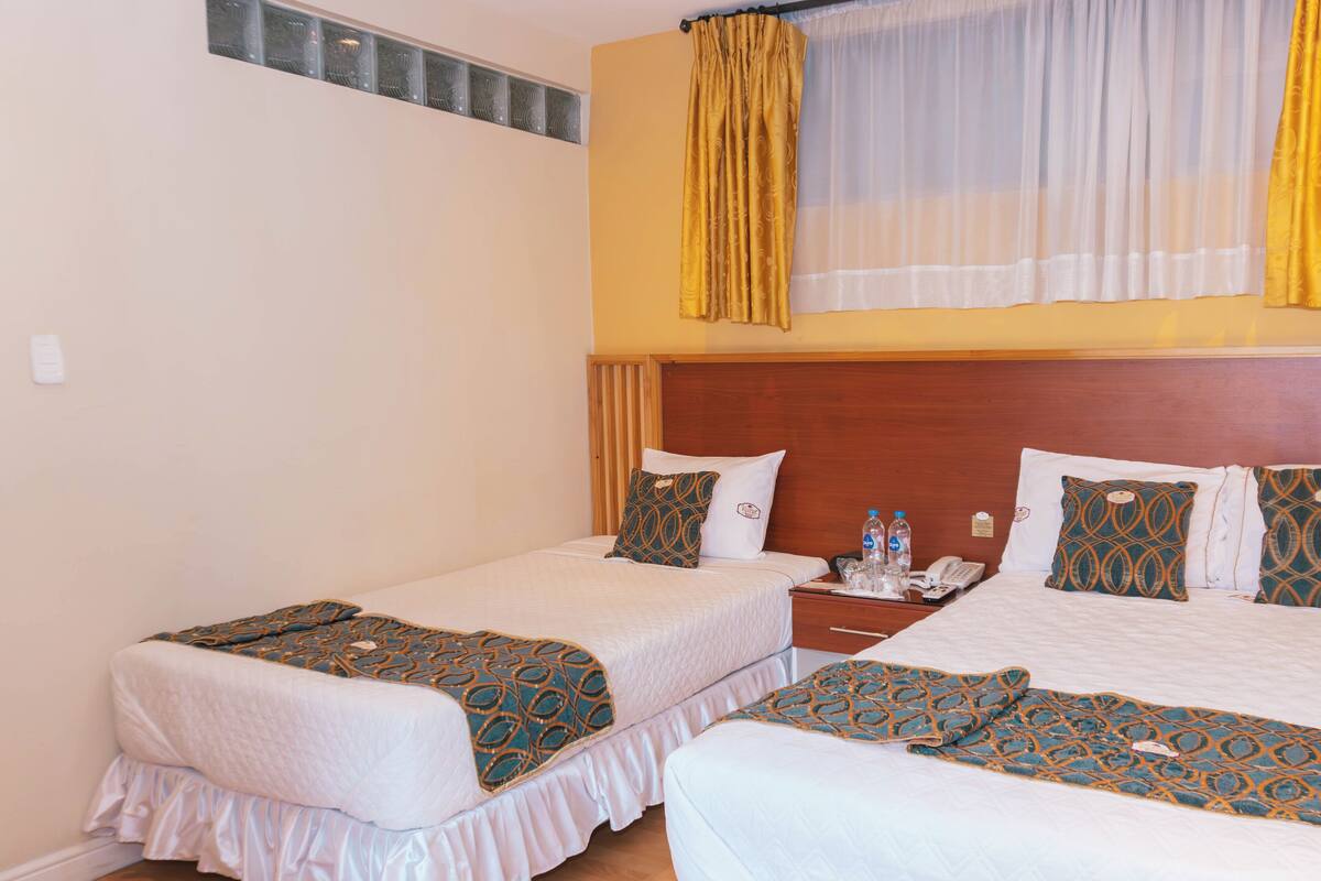 Habitación de hotel con dos camas, cabecero de madera, mesita de noche con un teléfono y botellas de agua, y ventana con cortinas amarillas.
