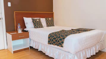 Una cama bien tendida con un plaid estampado, una mesita de madera con una botella y un control remoto, y un módulo de cajones blancos.
