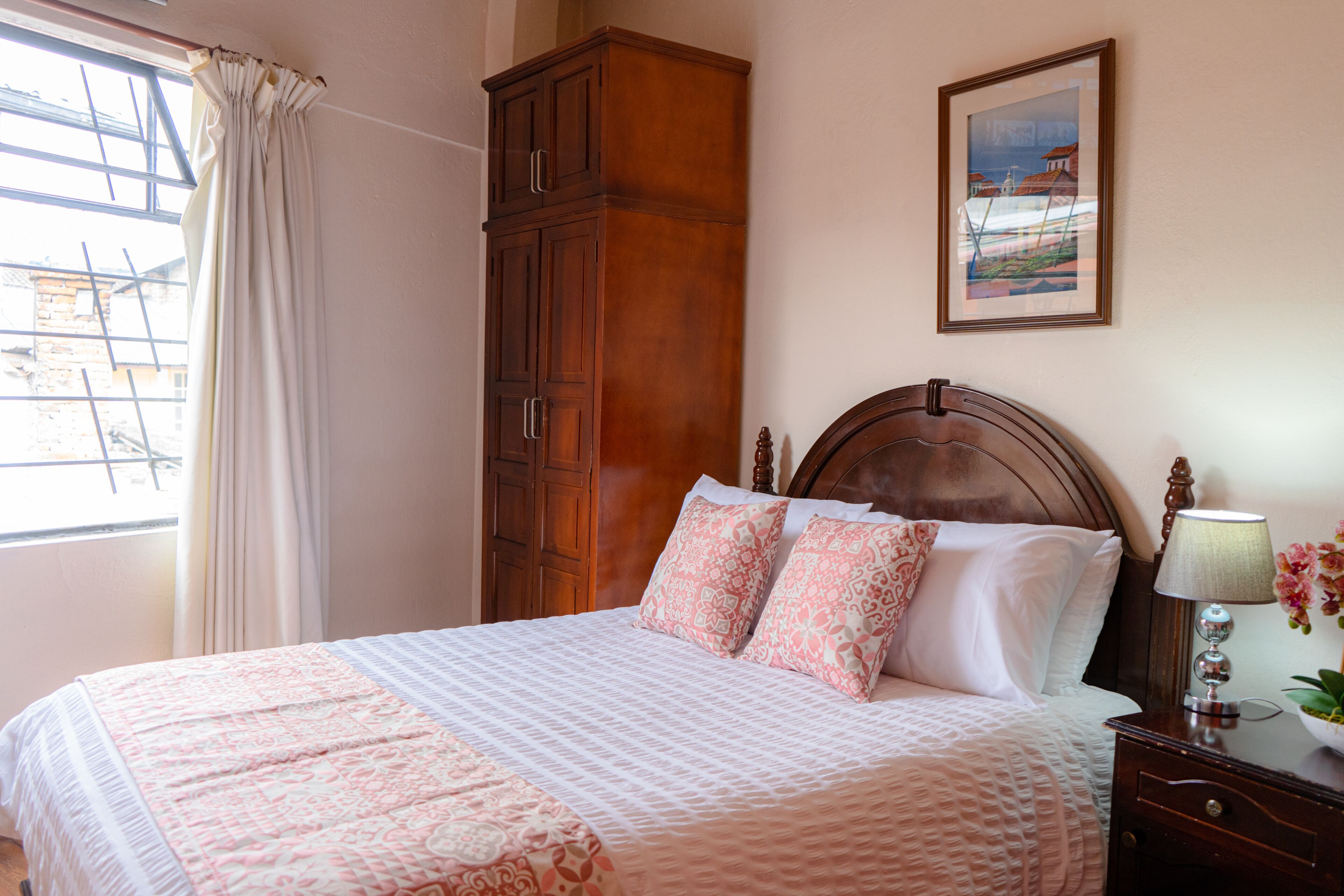 A neatly made bed with patterned bedding, a wooden wardrobe, a framed picture on the wall, and a bedside table with a lamp and flowers.