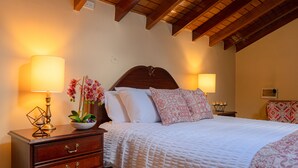 A neatly made bed with a white comforter and patterned pillows, a wooden nightstand with a lamp and a small plant, and a wooden dresser with drawers. - Rincón Familiar Hotel Boutique (Quito)