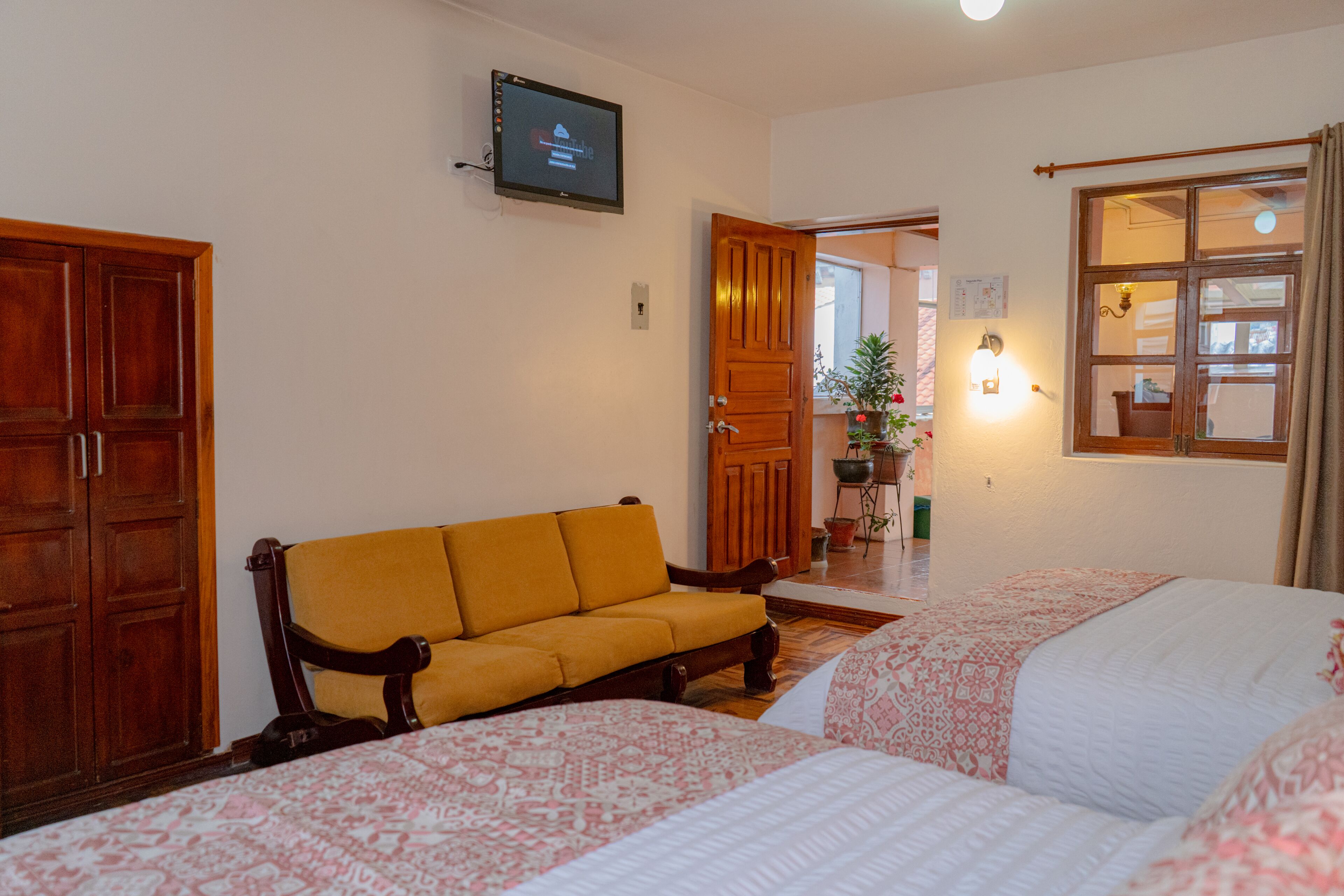 A hotel room with a wooden wardrobe, a sofa, two beds with patterned bedding, a television on the wall, and a window with curtains.