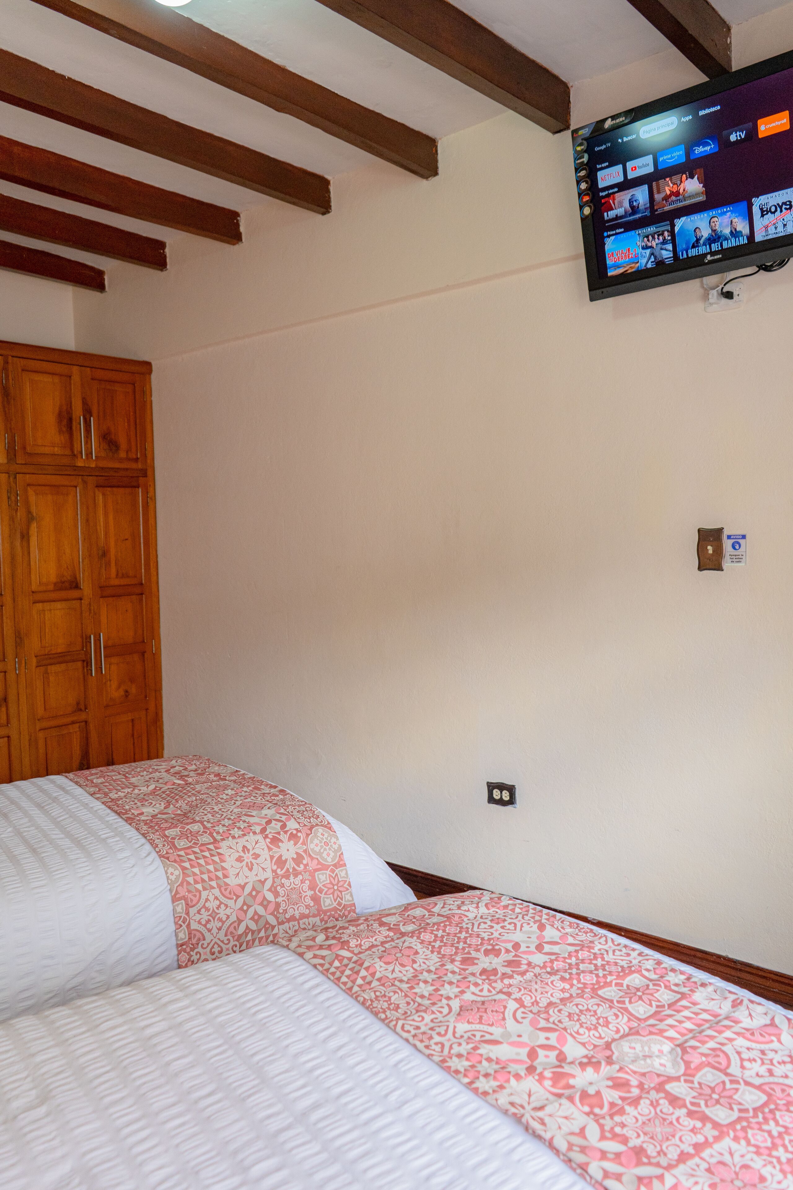 A bedroom with two beds, wooden wardrobe, and a mounted TV displaying various channels.