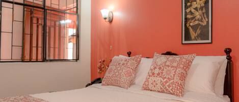 A bedroom with a bed, patterned pillows, a wall-mounted light, and a window with bars.