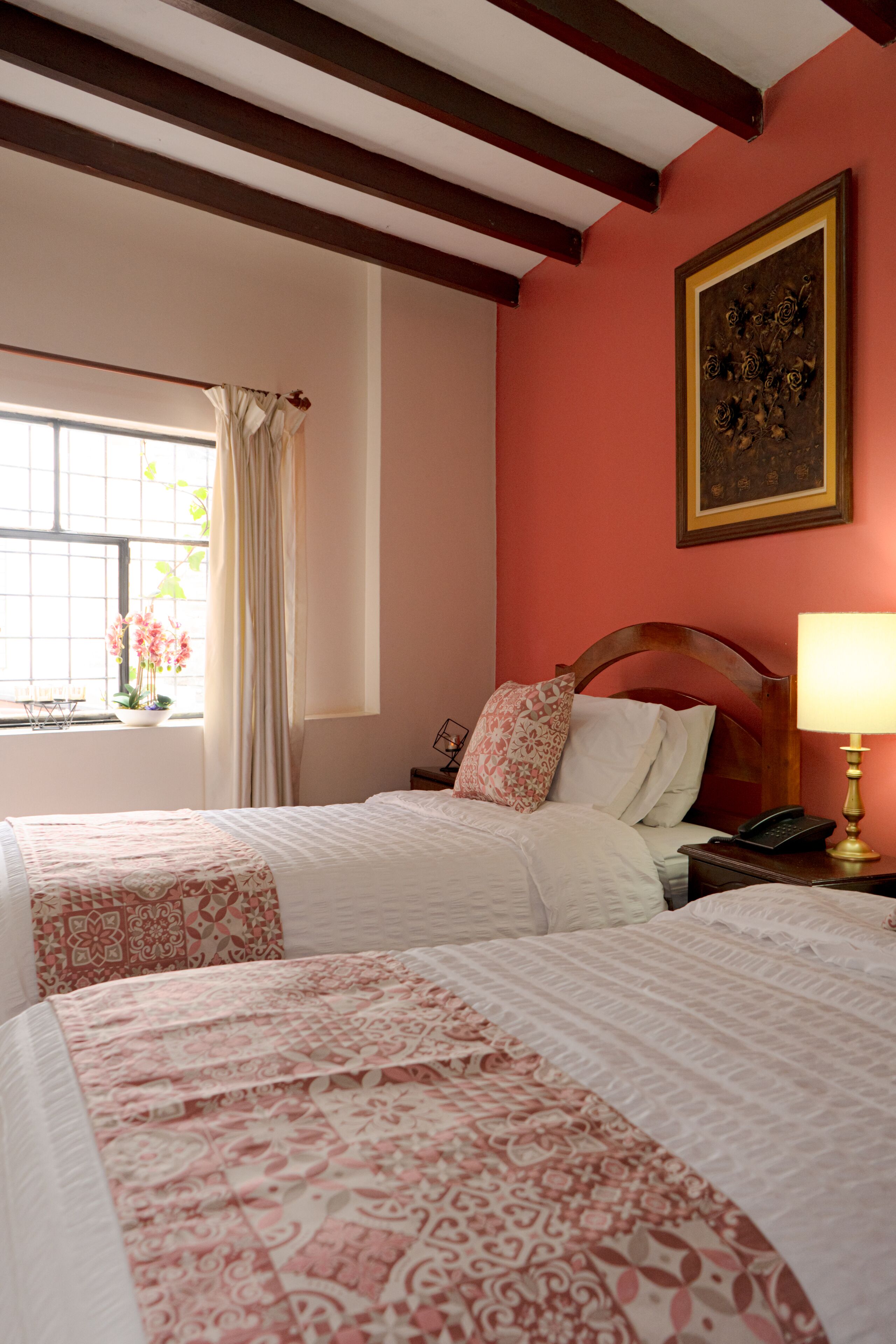 A hotel room with two beds, a window with curtains, a bedside table with a lamp, and a framed picture on the wall.