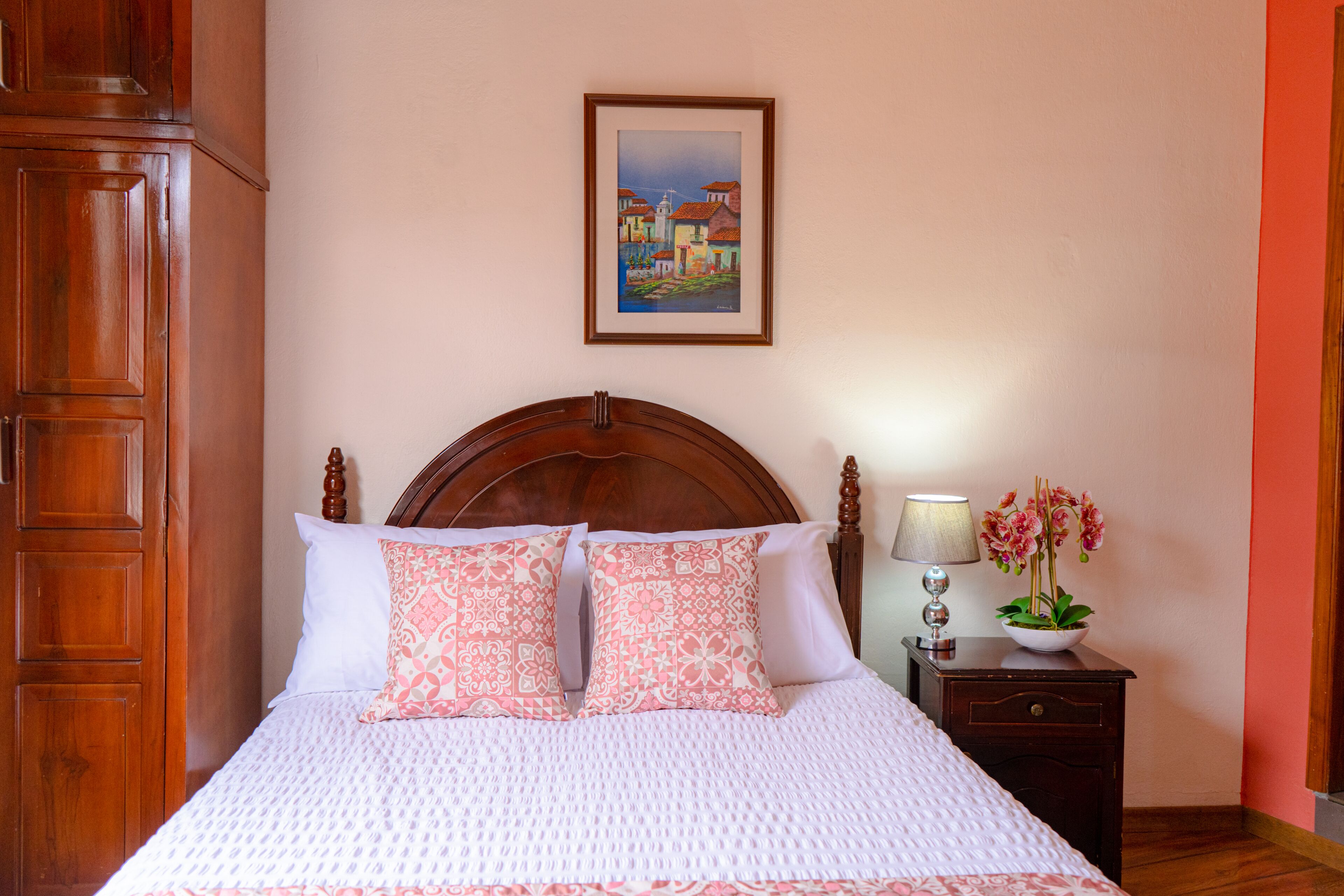 Una cama bien hecha con almohadones estampados, un cabecero de madera, una mesita de noche con una lámpara y flores, y un cuadro enmarcado en la pared.