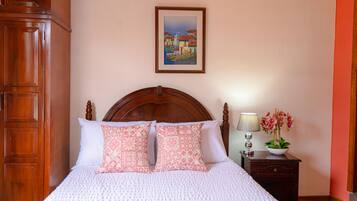A neatly made bed with patterned pillows, a wooden headboard, a bedside table with a lamp and flowers, and a framed picture on the wall.