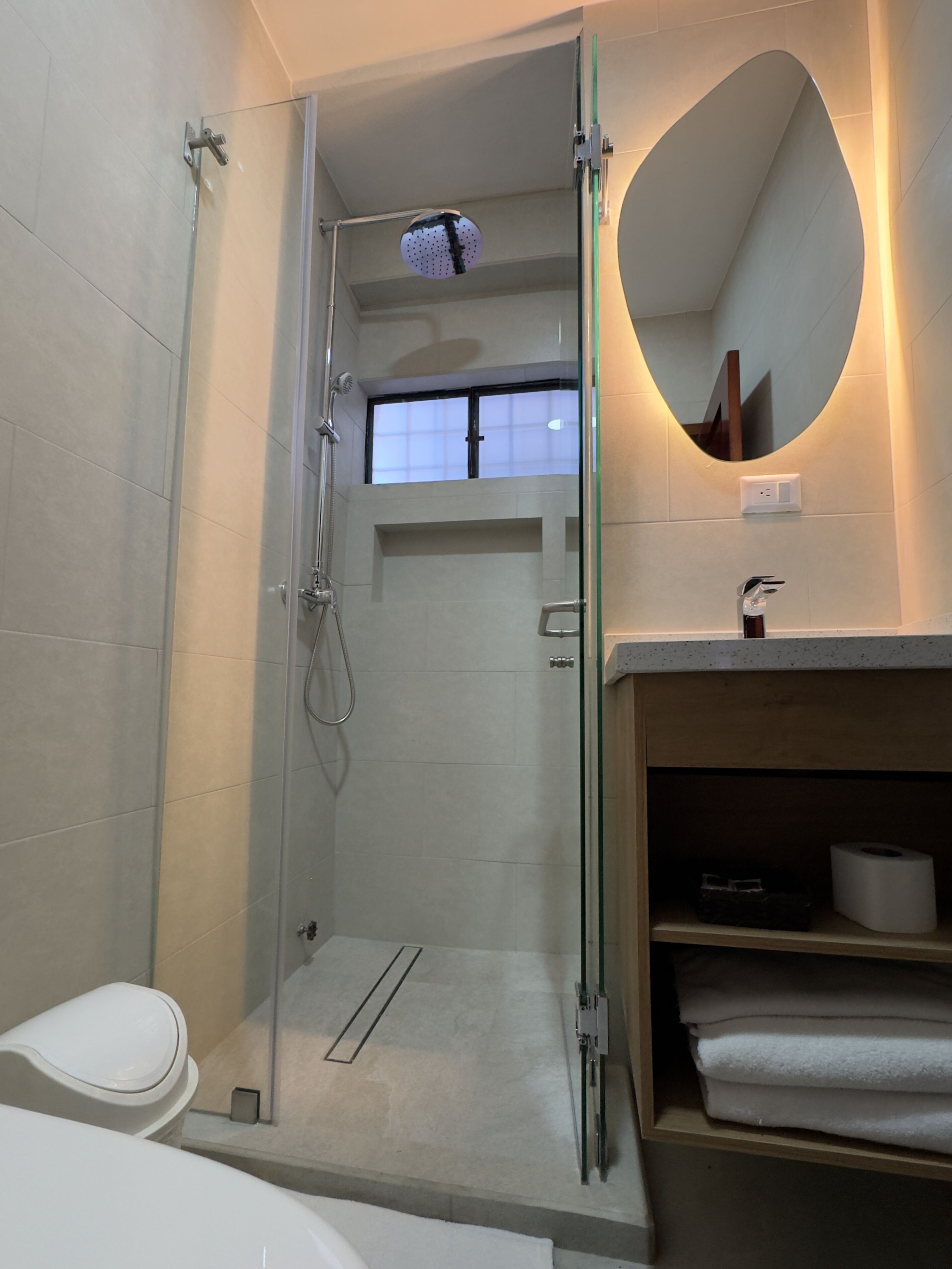 Un baño moderno con ducha de vidrio, inodoro, lavamanos con espejo y repisa con toallas.