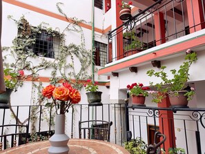 Une cour avec un sol carrelé, un vase blanc rempli de fleurs et des plantes en pot disposées sur la rambarde d’un balcon.