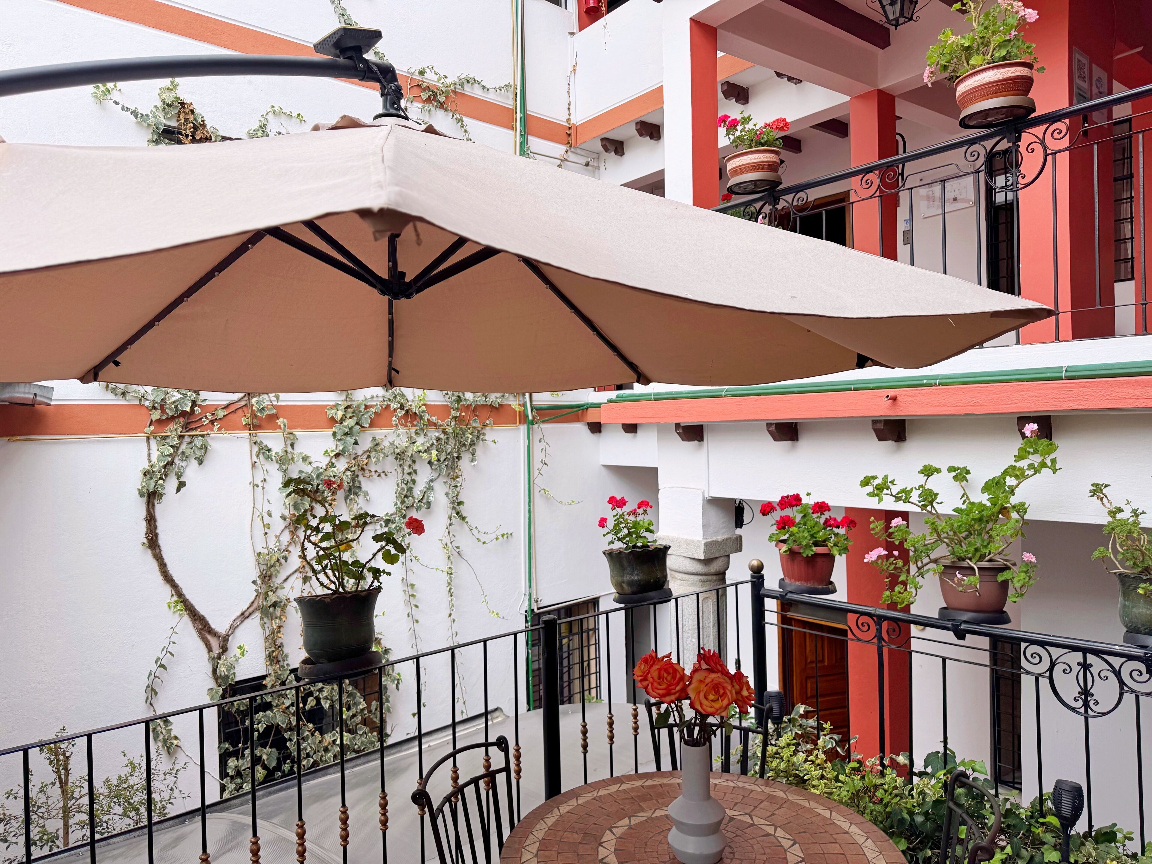 A covered outdoor seating area with a table and chairs, surrounded by potted plants and a railing.