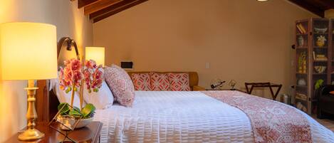 Quarto com cama grande, mesa de cabeceira com abajur, cabeceira de madeira, mesinha de cabeceira com abajur e uma planta decorativa.