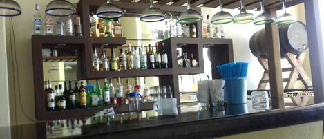 Zona de bar con estantes llenos de botellas de licores, lámparas colgantes y un barril de madera.