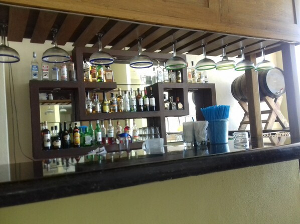 Zona de bar con estantes llenos de botellas de licores, lámparas colgantes y un barril de madera.