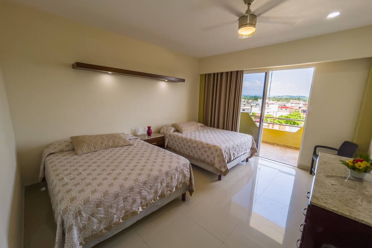 A hotel room with two beds, a ceiling fan, a nightstand, a desk, and a view of the city.