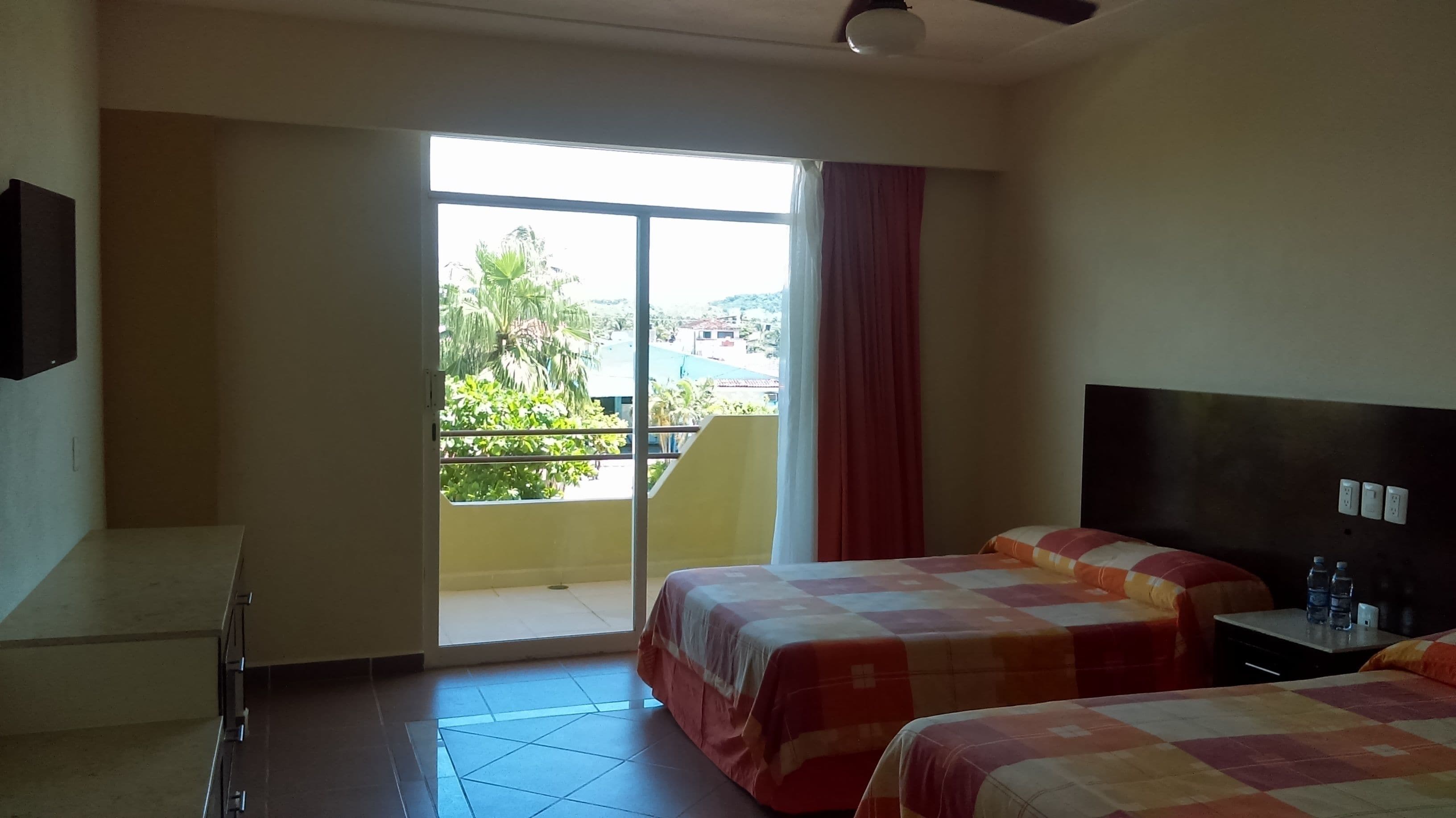 A hotel room with two beds, a desk, a chair, a window with a view of greenery and buildings, and a ceiling fan.
