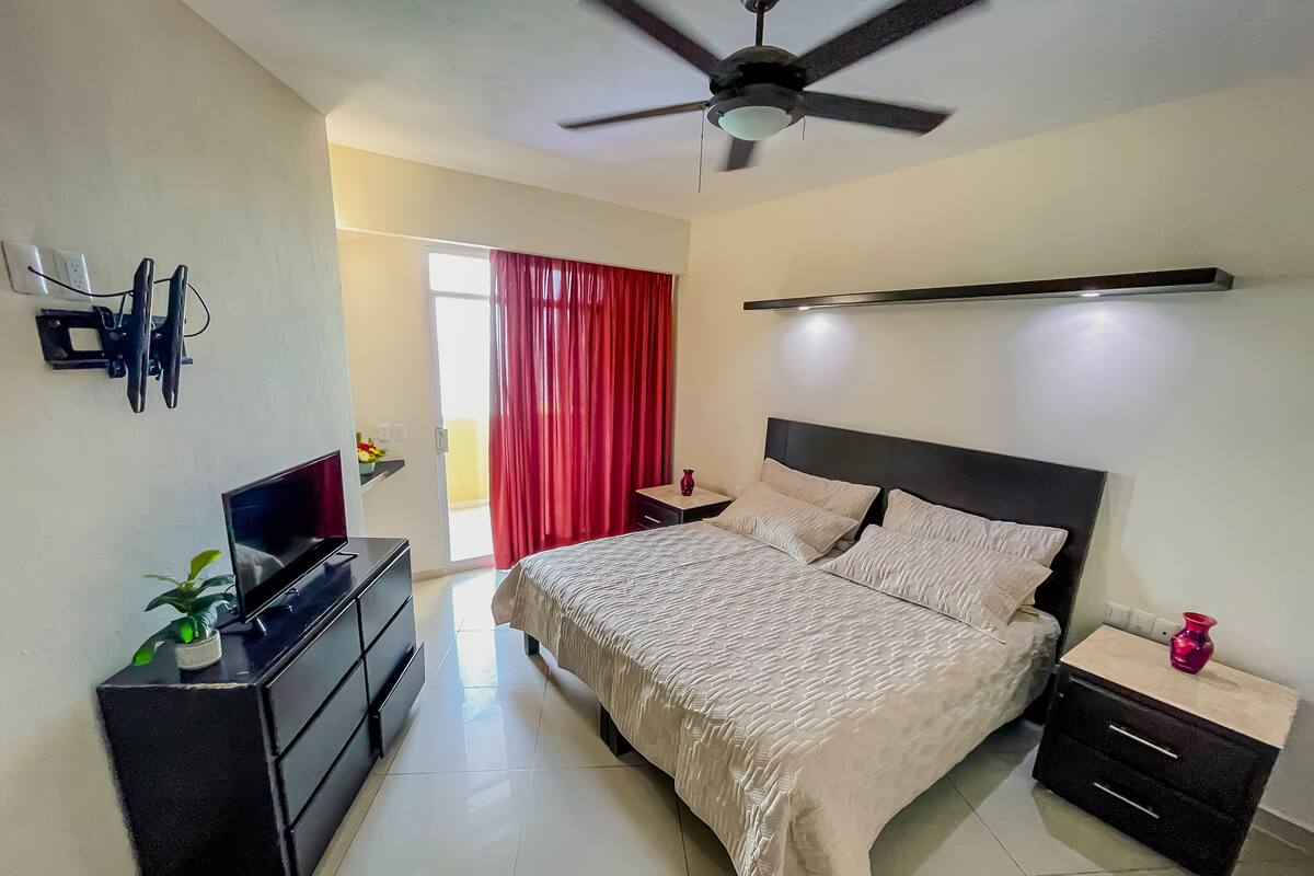 A bedroom with a bed, bedside tables, a TV, a ceiling fan, and a red curtain.