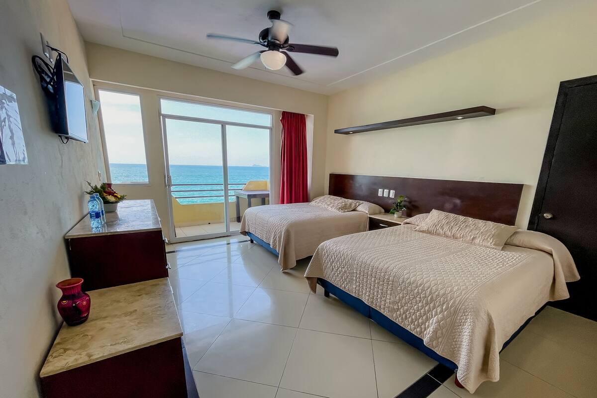 A hotel room with two beds, a ceiling fan, a desk with a chair, a small table with a vase, and a view of the sea through a sliding door.