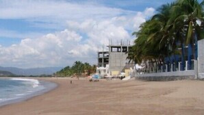 A beach with a building under construction, palm trees, and mountains in the background. - Hotel Bahía (San Patricio Melaque)