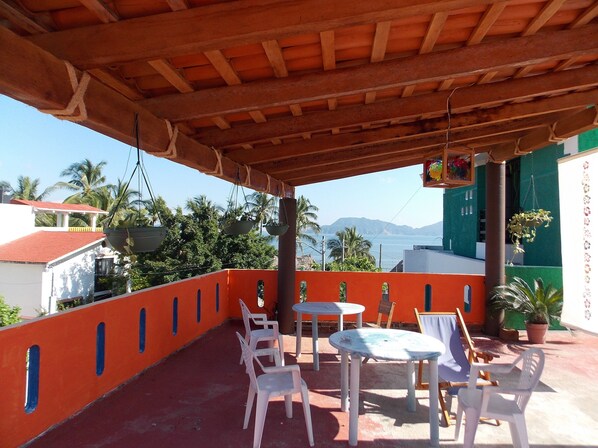 A covered patio with white plastic chairs and tables, overlooking a coastal view with palm trees and a mountain in the distance.