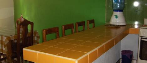 A kitchen with a long table, chairs, a blender, and a trash bin.