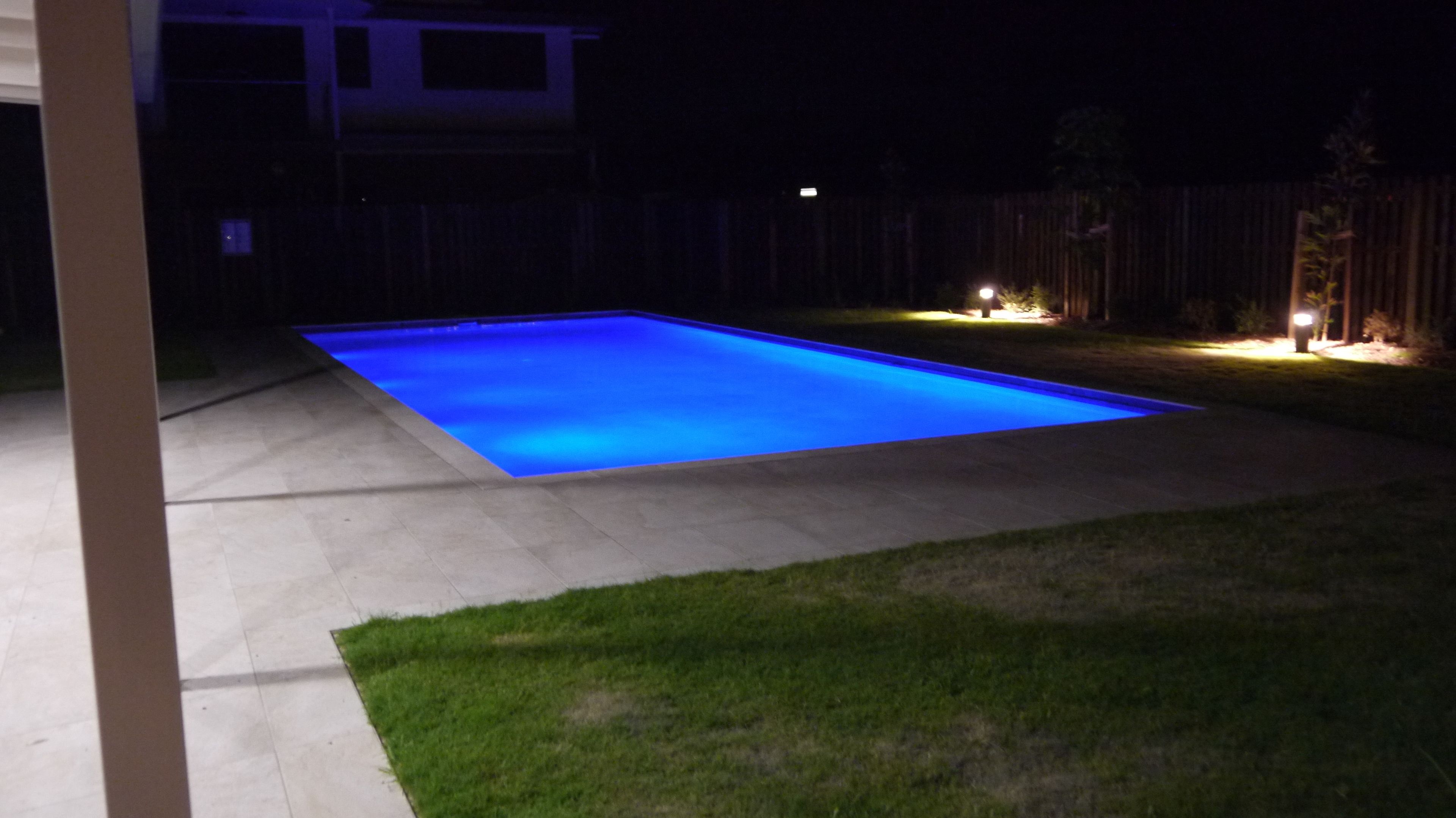 Une piscine rectangulaire dont le bord est illuminé en bleu, entourée d’une terrasse en béton et d’une zone herbeuse.