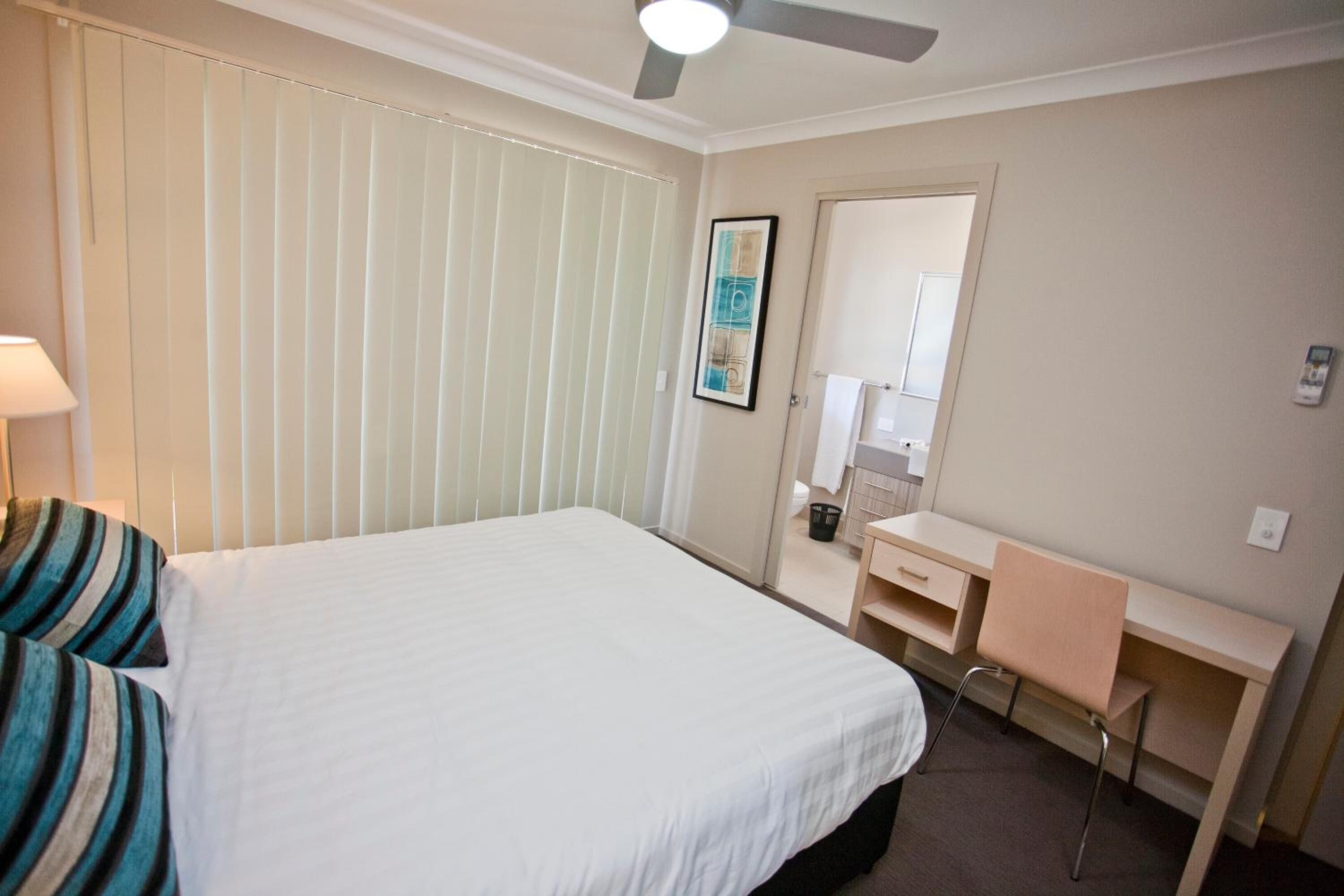 A hotel room with a bed, a desk, a chair, a ceiling fan, and a window with blinds.