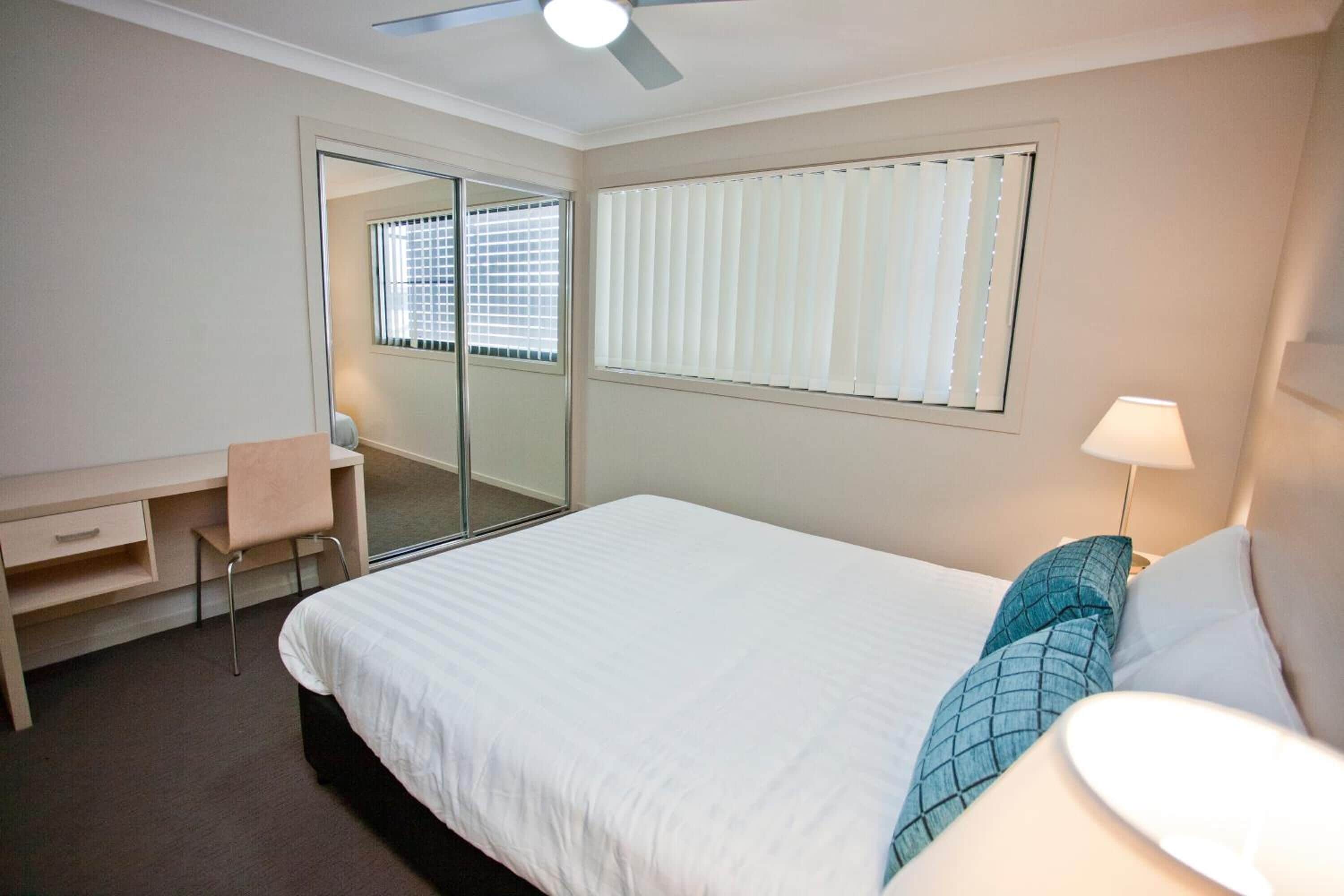 A bedroom with a bed, a desk, a chair, a lamp, and a window with blinds.